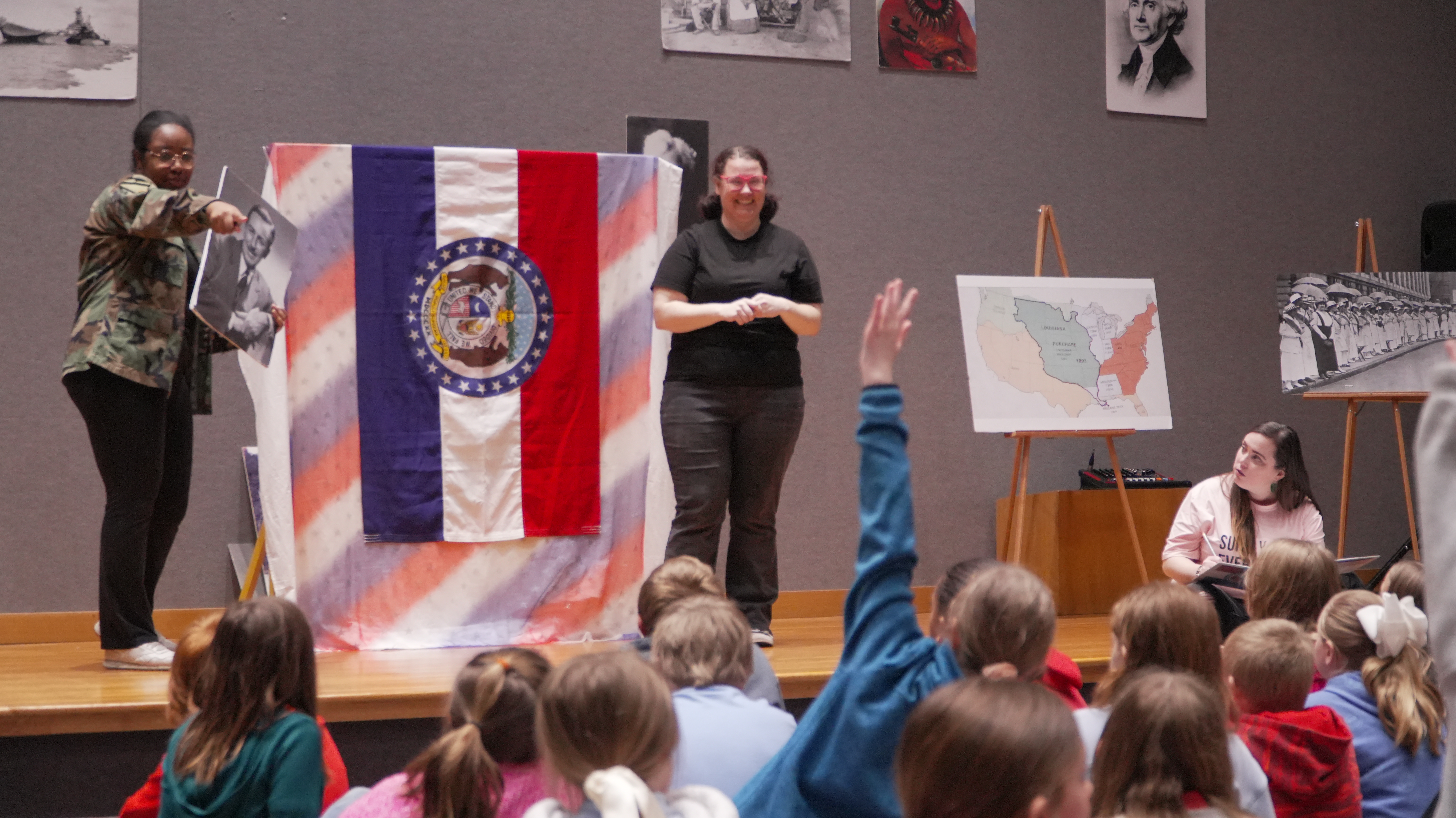 Color image of Actresses performing on stage and holding up images of famous missourians for an audience of young students.